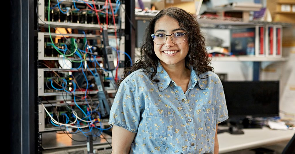 Female student smiling. She is standing in front of a bank of main fame computers.
