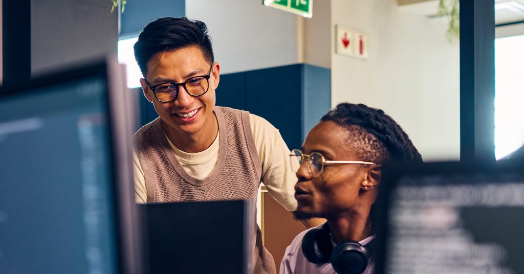 A lecturer standing by a seated student discussing his screen's content.