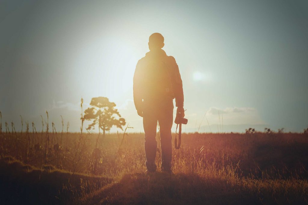 A man is standing in a field, back to the reader. He holds a camera by his side. It's important we develop an understanding of why the arts are important to our regions.