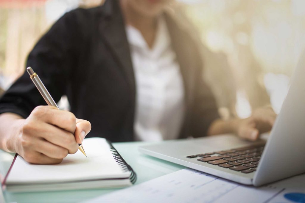 Close up of woman working on a laptop while writing on a notepad.