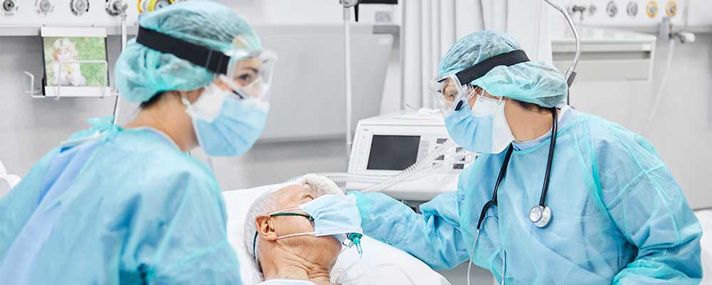 Two healthcare workers in full PPE, caring for an elderly man on a ventilator 