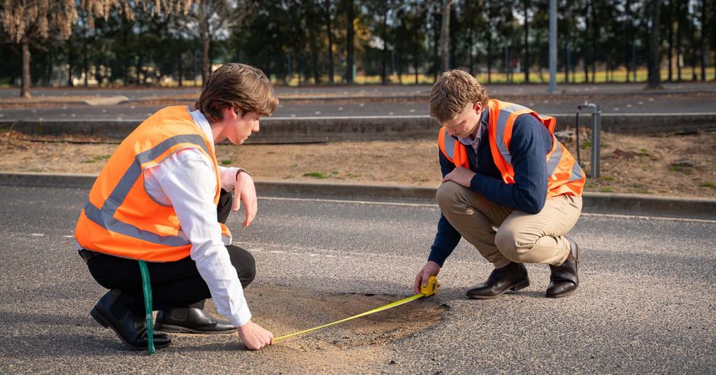 Two male engineers in high vis vests use a tape measure to measure a pothole on the road