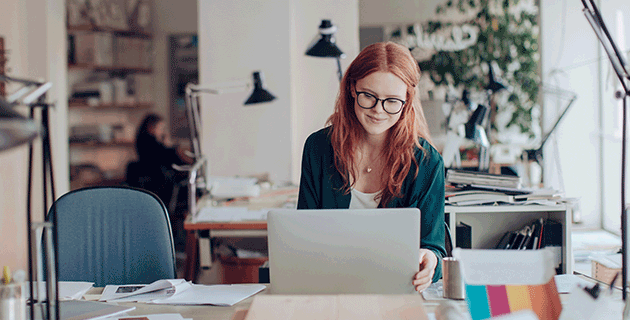 A scene featuring a marketing manager (a key Bachelor of Communication job) with red hair and glasses seated at a collaborative and creative workspace, typing on her laptop.