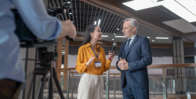 A smiling female journalist, who's wearing a yellow top and beige suit pants, interviewing a man with a blue suit and tie on. 