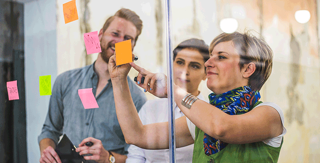 A woman in an advertising workplace, standing while writing on a post-it note, two team members in the background collaborate, capturing idea generation and teamwork in a Bachelor of Communication job.