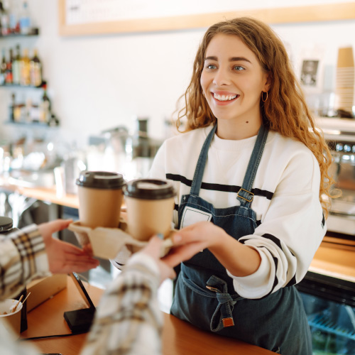 A person stands behind a counter and hands over a set of two coffees that a customer has ordered.