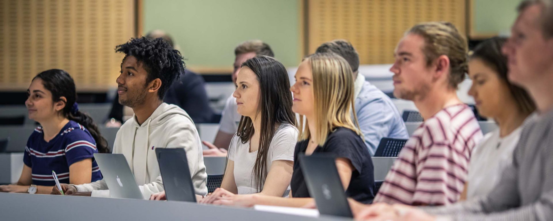 A bunch of male and female students sit in a lecture hall, typing notes on their laptops