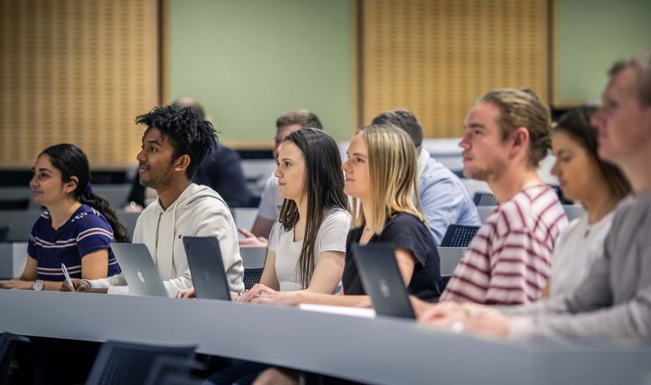 A bunch of male and female students sit in a lecture hall, typing notes on their laptops