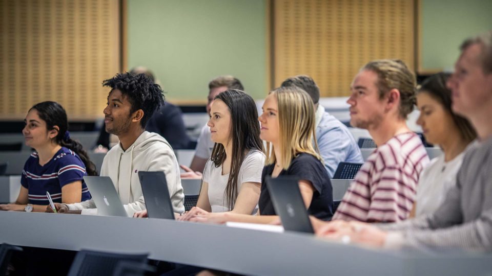 A bunch of male and female students sit in a lecture hall, typing notes on their laptops