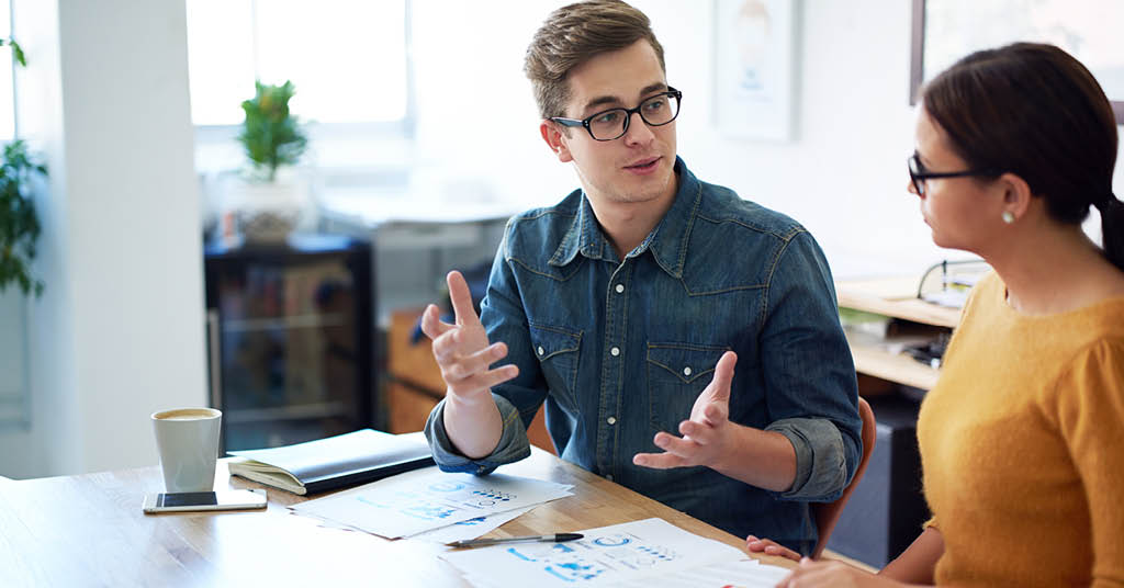 A male and female colleague discuss work in an office environment, conveying communication and collaboration in a human resources context.