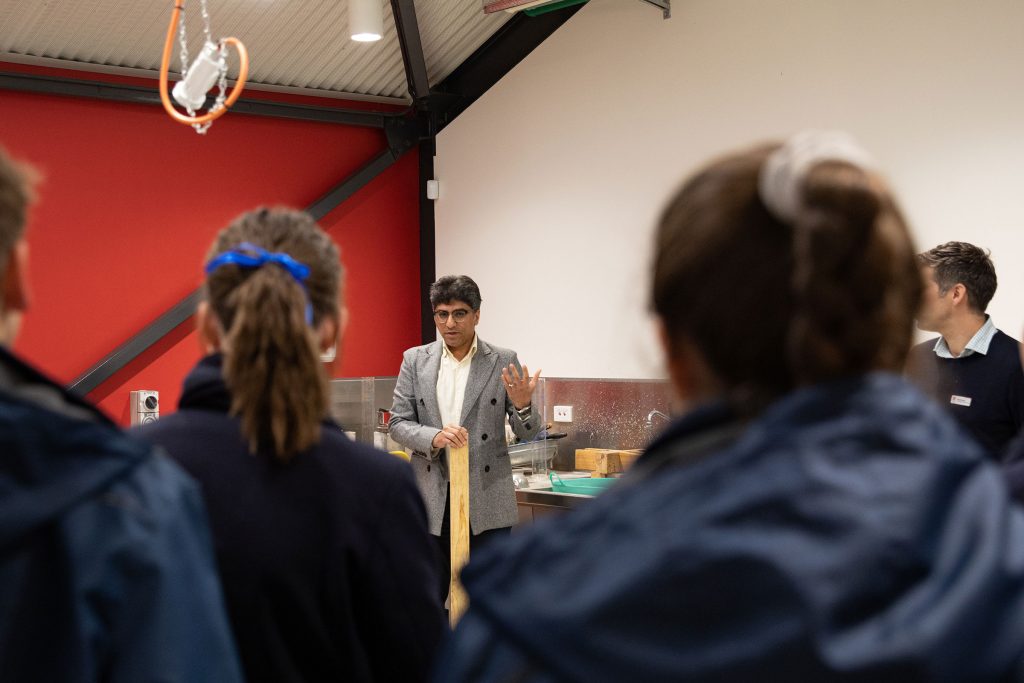 A lecturer in a grey coat holds a piece of wood as he teaches some students about engineering.