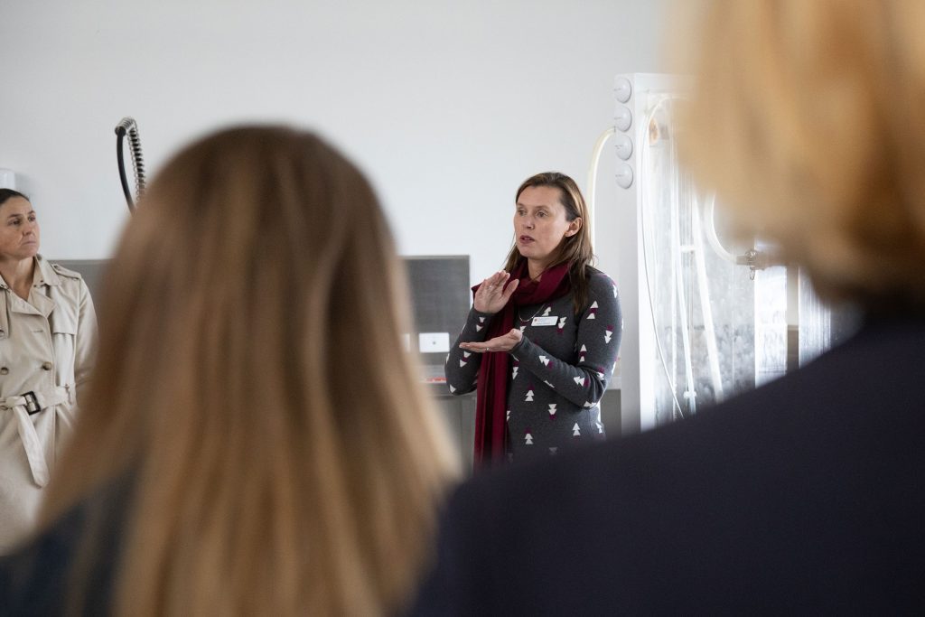 A lecturer in a red scarf and patterned grey sweater is indicating size with her hands as she teaches students in a lab.