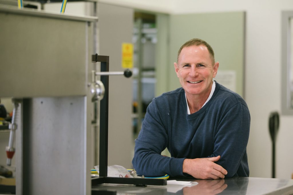 A lecturer in a grey pullover sweater smiles as he sits inside a lab.