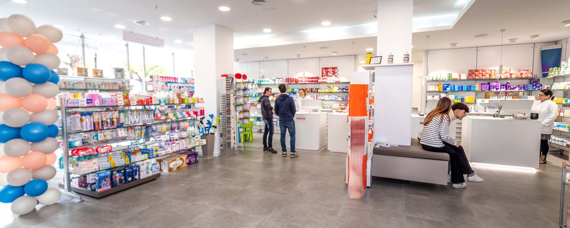 Two people buying items at a pharmacy. Female cashier in white coat serves customer. Shelves with health care products.