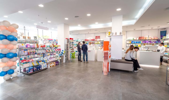 Two people buying items at a pharmacy. Female cashier in white coat serves customer. Shelves with health care products.