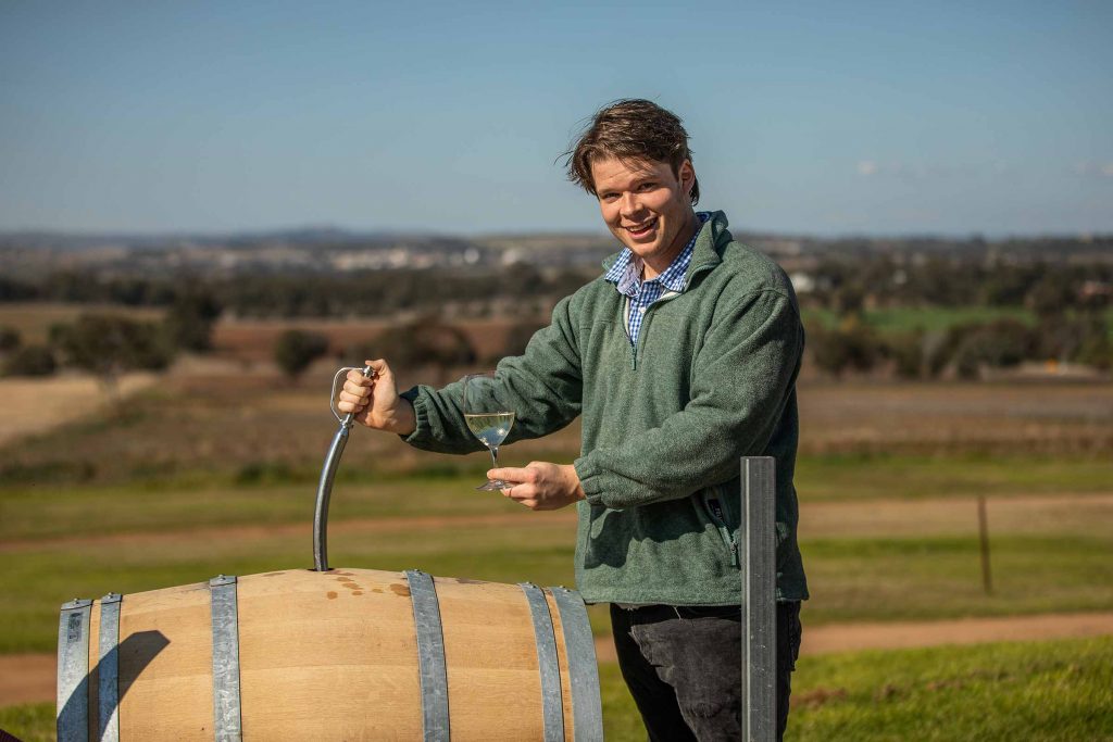 
A young man, smiling at the camera, is pouring white wine from a barrel into a glass. 