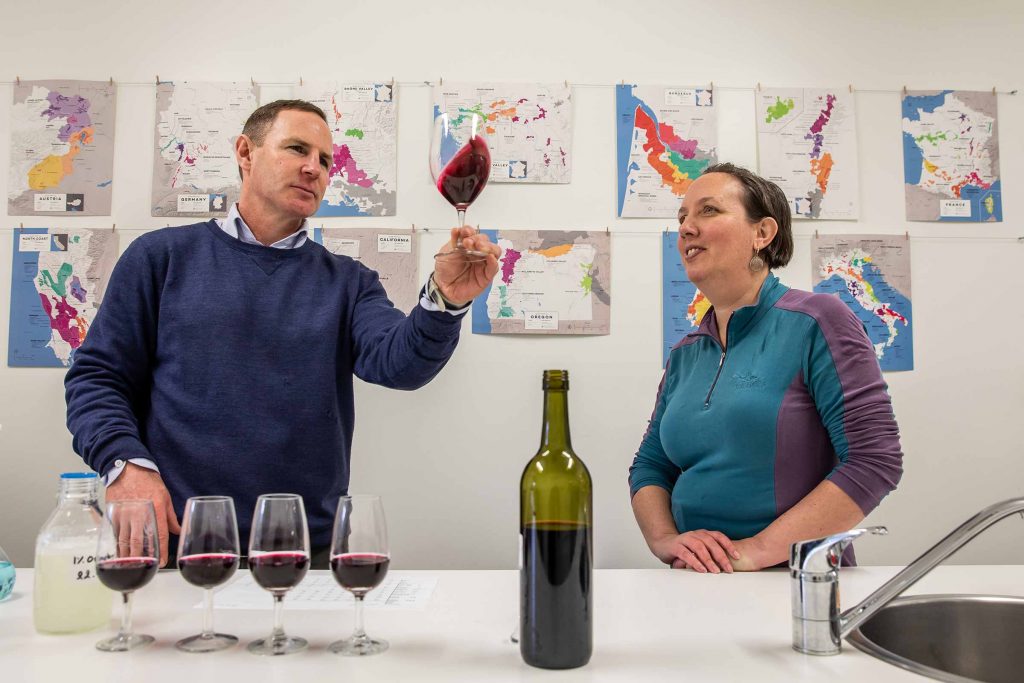 A man, positioned to the left, holds a glass of red wine and swirls it, examining its qualities, while a woman stands beside him, observing his actions. Four glasses of red wine are placed on a table, suggesting they are testing or evaluating the wine together.