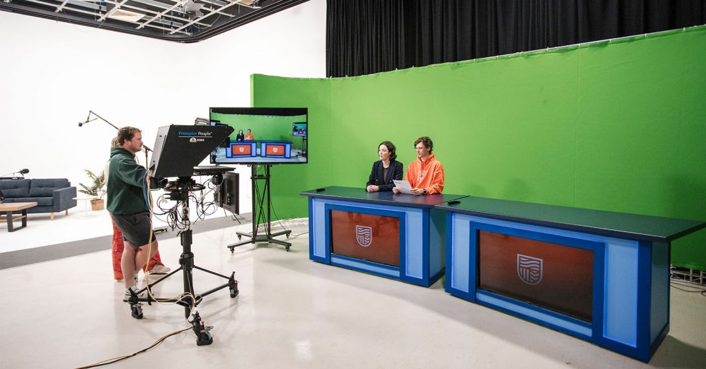 Students in the Charles Sturt news room presenting at a professional news desk in front of a green screen, showcasing the media precinct’s industry-standard setup.