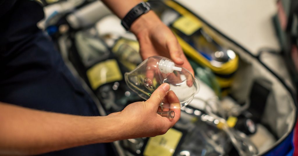 A student paramedic's hands holding an oxygen mask, with a simulation ambulance kit in the background