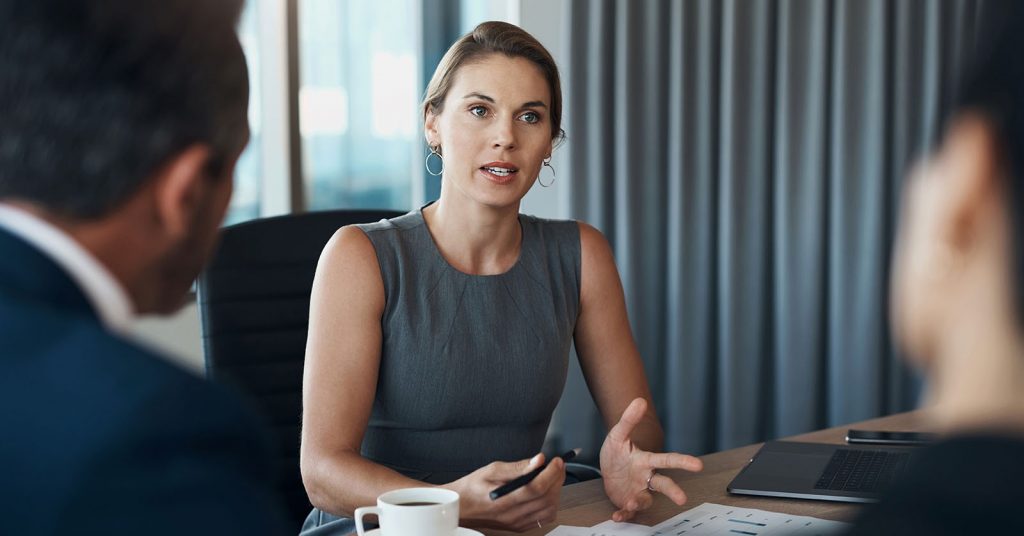 A businesswoman speaks with two colleagues during a meeting, seated around a conference table in a modern meeting room, representing collaboration and teamwork in a professional setting.
