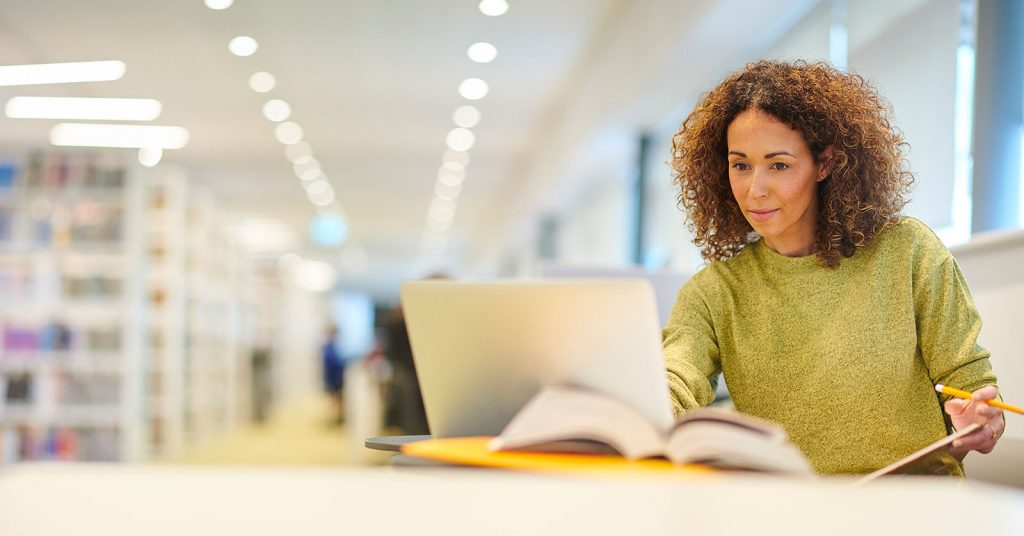 A woman studies on a laptop in a quiet library, surrounded by bookshelves, illustrating focused learning in an academic environment.