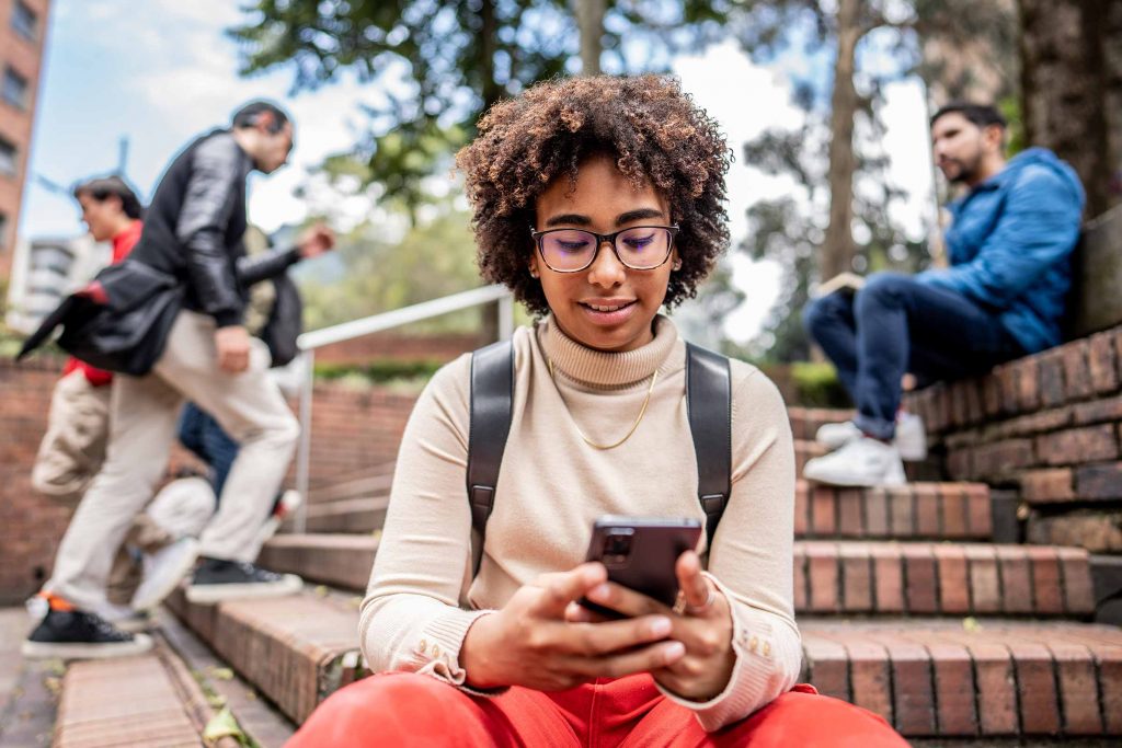 A girl holding and looking at a mobile phone, with three men walking up steps in the background and one man sitting on the step.