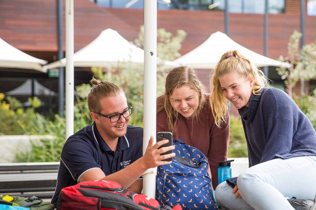 Three university students smiling and looking at a mobile phone together on a campus. 