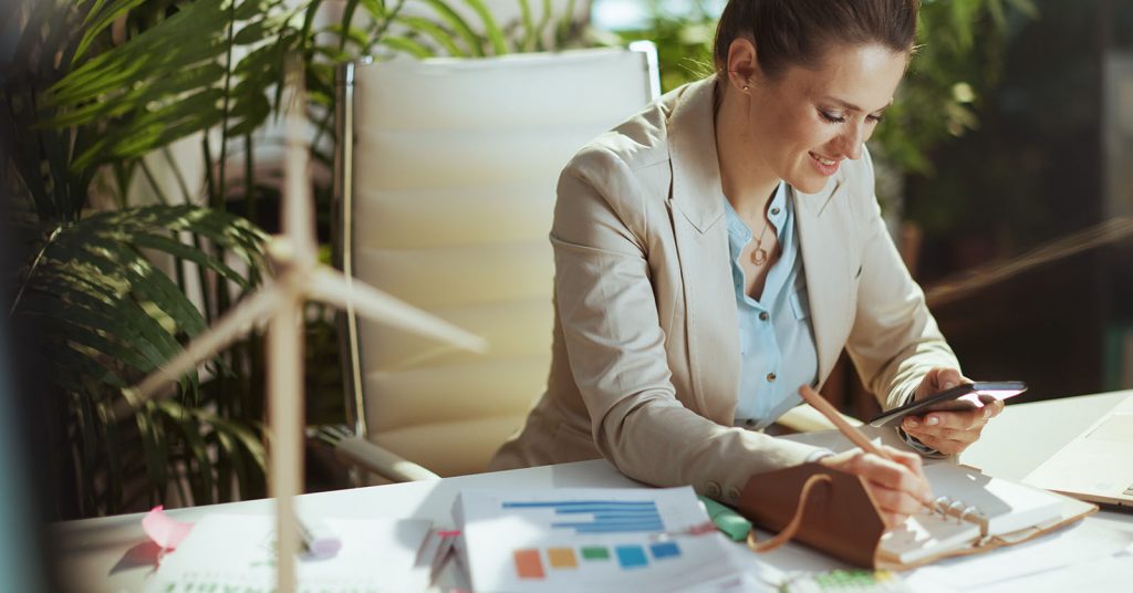 A woman writing in a notebook while looking at her phone