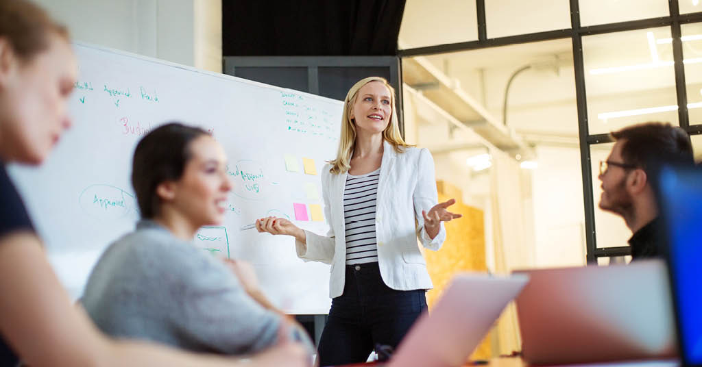 A woman with her arms open with a whiteboard behind her talking to a class of students 