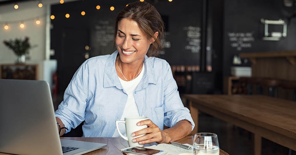 A woman holding a coffee mug, smiling and looking at a laptop