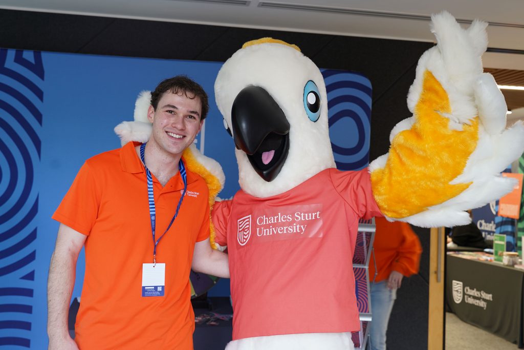 A male student with Charlie the cockatoo mascot smiling for the camera with a blue Charles Sturt banner in the background. 