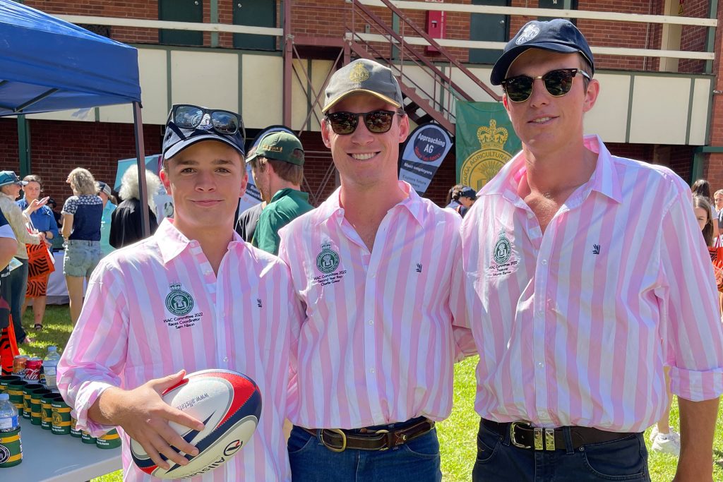 Three men, all wearing sunglasses and identical striped pink shirts, with one holding a rugby ball.