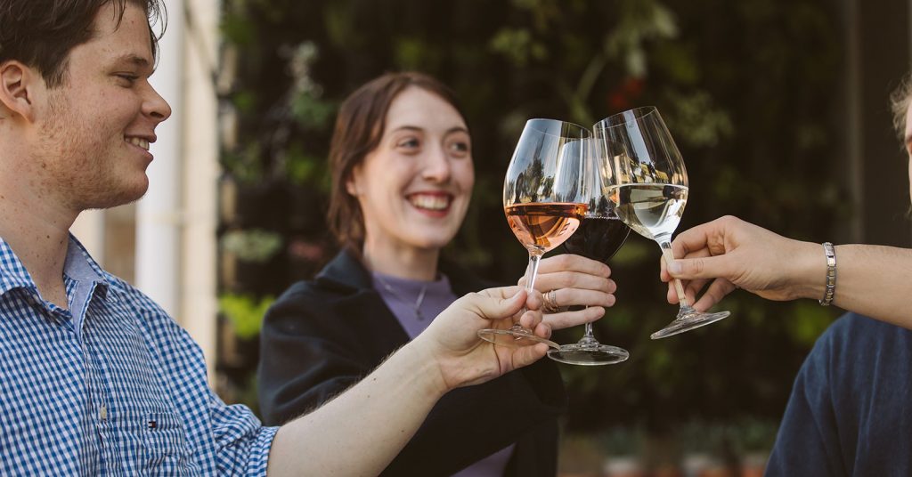 A group of three young people are cheerfully toasting with glasses of wine. Each person holds a different type of wine—one with white wine, another with rosé, and the third with red wine—celebrating together.