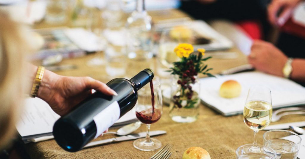 A person is pouring red wine into a glass at a restaurant, carefully filling it from a bottle. The setting appears elegant, with a focus on the wine being served.