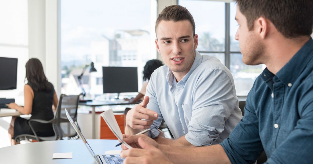 Two men chatting at a desk, with a laptop in front of them