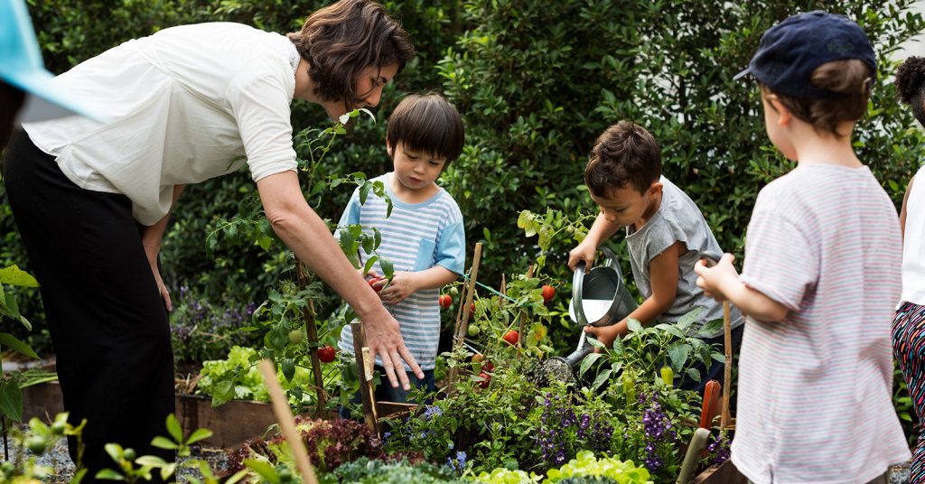 A teacher guiding young children as they plant vegetables in a school garden, demonstrating hands-on learning and inclusion through shared participation.