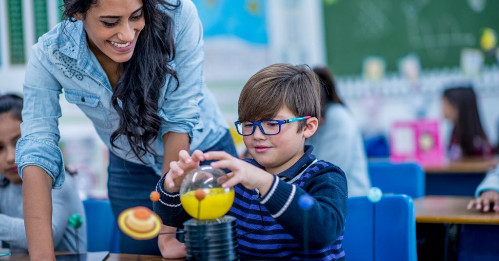 A smiling teacher guides a student through a hands-on science lesson, demonstrating practical, engaging learning in the classroom.