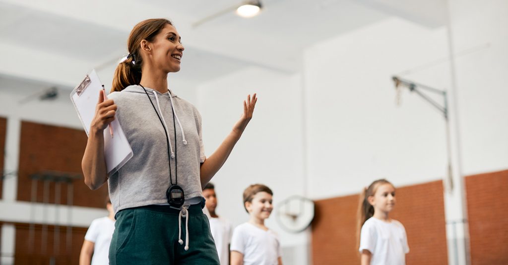 A smiling physical education teacher stands on a basketball court with students lined up in the background, representing active learning and inclusive participation in sport.