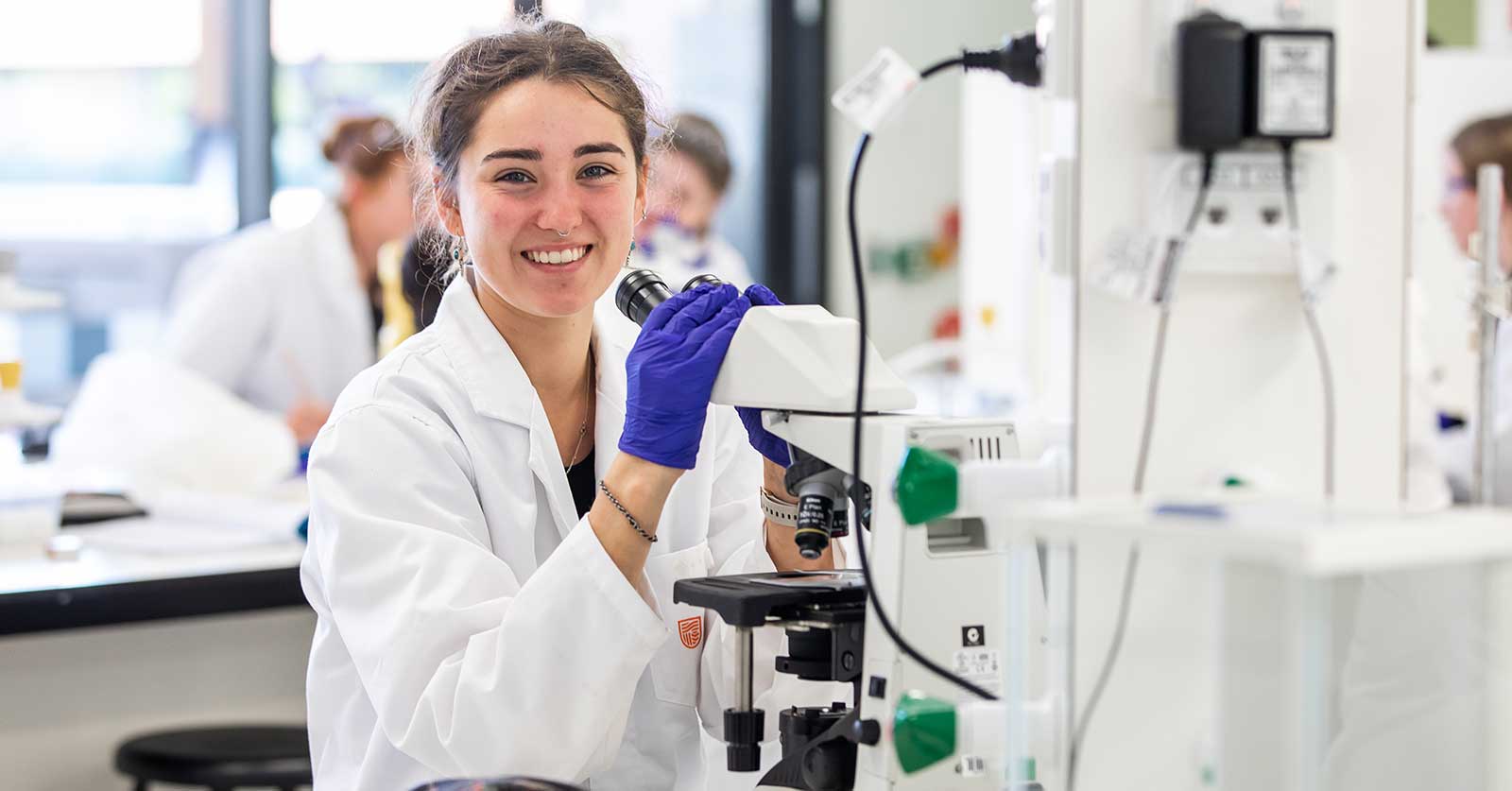 young lady smiling and holding onto a microscope in a pathology lab