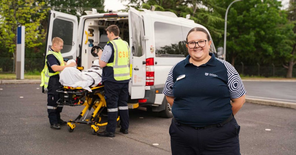Female paramedicine student smiling in the foreground while other students conduct an emergency simulation, loading a simulated patient into a Charles Sturt University ambulance in the background.