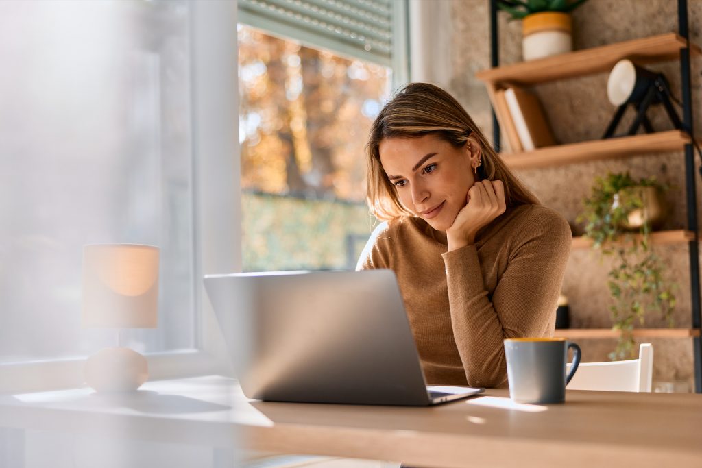 A young person with long blonde hair sits at a light wooden desk, looking thoughtfully at a laptop screen. She is wearing a brown long-sleeved sweater and rests her chin on her hand. A blue ceramic mug sits on the desk beside her laptop, and a small white lamp is visible in the foreground. Behind her, a large window allows soft, natural light to fill the room, showing a glimpse of autumn trees outside. To the right, a wooden shelving unit holds books and a small potted plant.