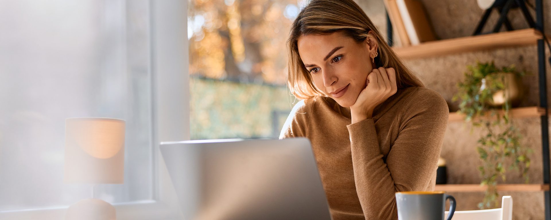 A young person with long blonde hair sits at a light wooden desk, looking thoughtfully at a laptop screen. She is wearing a brown long-sleeved sweater and rests her chin on her hand. A blue ceramic mug sits on the desk beside her laptop, and a small white lamp is visible in the foreground. Behind her, a large window allows soft, natural light to fill the room, showing a glimpse of autumn trees outside. To the right, a wooden shelving unit holds books and a small potted plant.