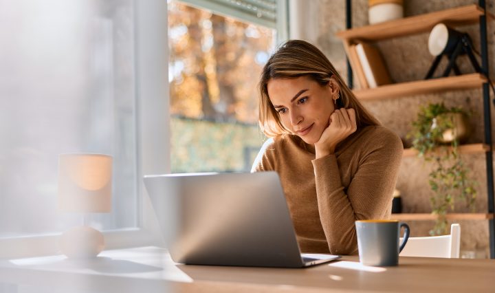 A young person with long blonde hair sits at a light wooden desk, looking thoughtfully at a laptop screen. She is wearing a brown long-sleeved sweater and rests her chin on her hand. A blue ceramic mug sits on the desk beside her laptop, and a small white lamp is visible in the foreground. Behind her, a large window allows soft, natural light to fill the room, showing a glimpse of autumn trees outside. To the right, a wooden shelving unit holds books and a small potted plant.