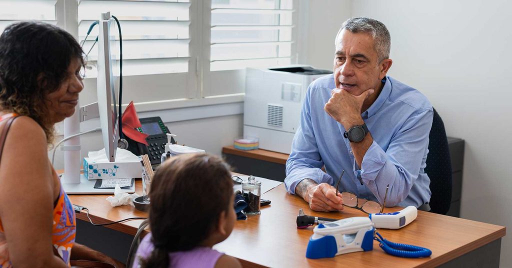 An older male doctor sits in a medical office speaking with a woman and a young girl, who are seated across from him during a consultation.