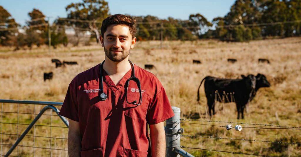 A man in his mid‑20s wearing medical student attire stands on a rural farm, with cows grazing in a paddock behind him.